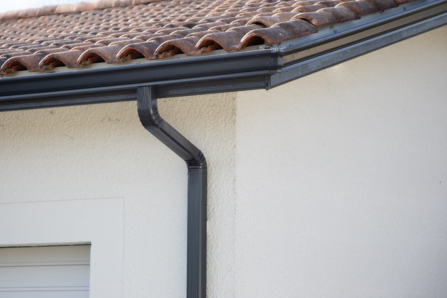 Close-up of a house corner showing a tiled roof, black seamless gutter system, and downspout on a stucco wall.