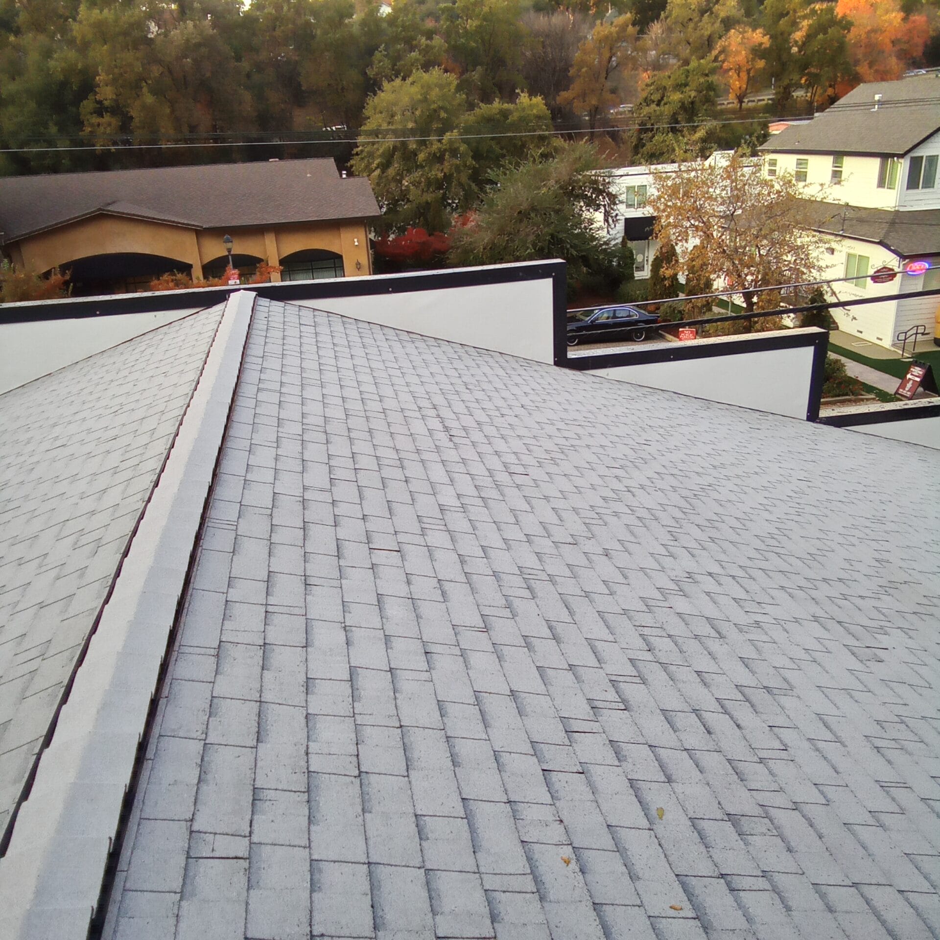 Close-up view of a gray commercial roof with shingle tiles, showcasing a clean roof ridge and surrounding autumn trees and buildings in the background.