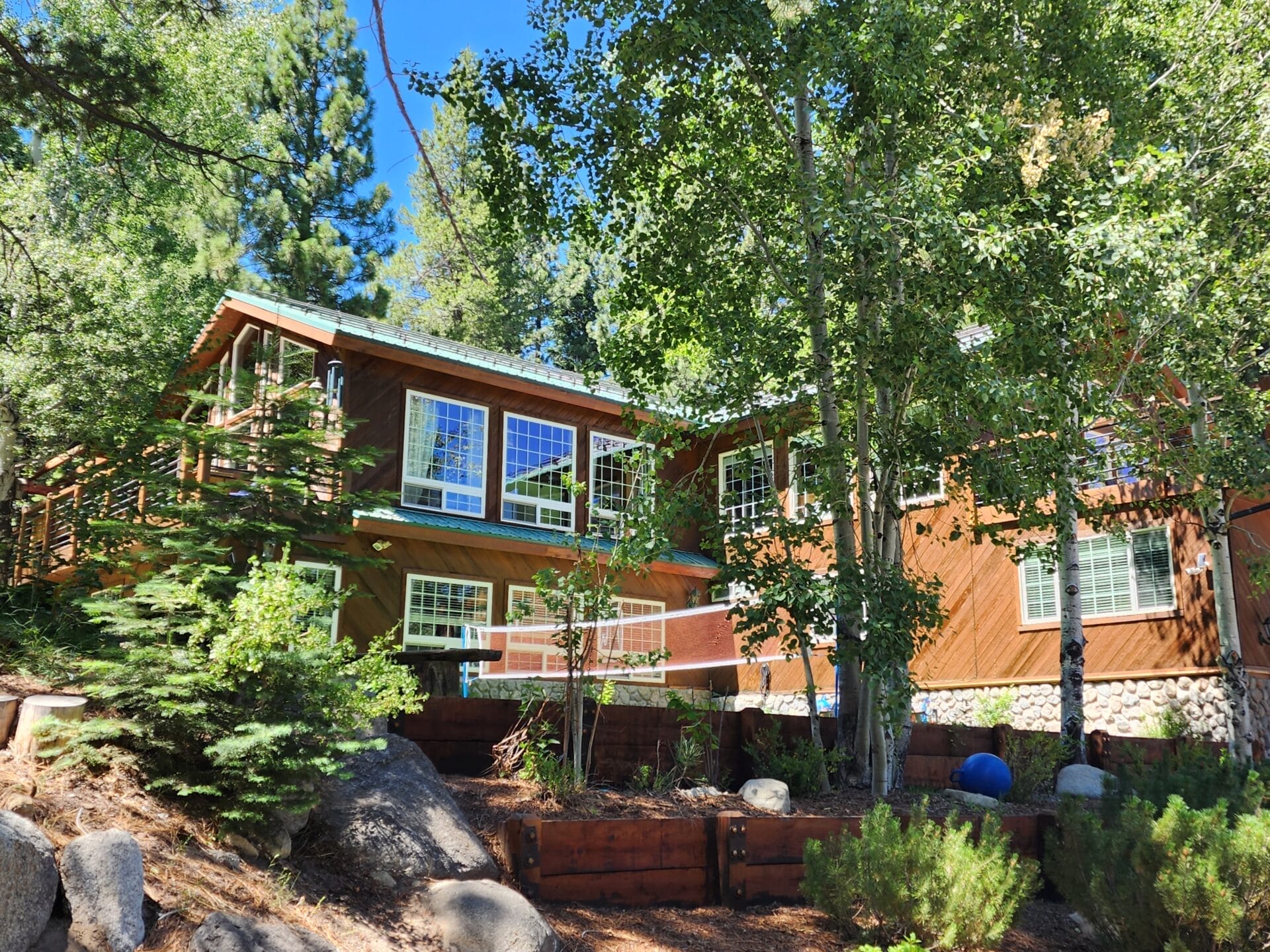 Two-story wooden house with large windows surrounded by tall trees and greenery