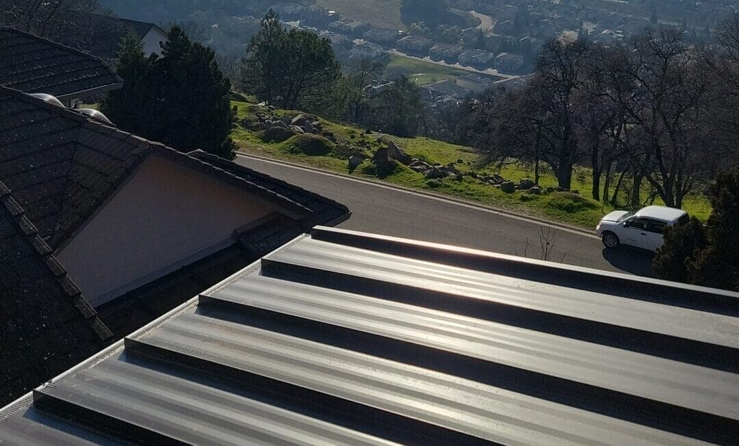 A sunny, scenic view of a vast valley and distant city, captured from a high vantage point. In the foreground is a corrugated metal roof.
