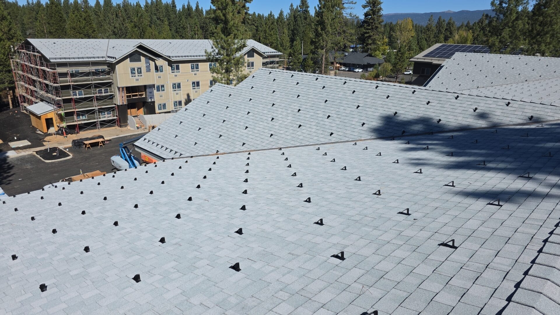 View from a rooftop covered in light gray shingles with many evenly spaced black mounts; construction site and apartment buildings surrounded by trees are visible in the background.