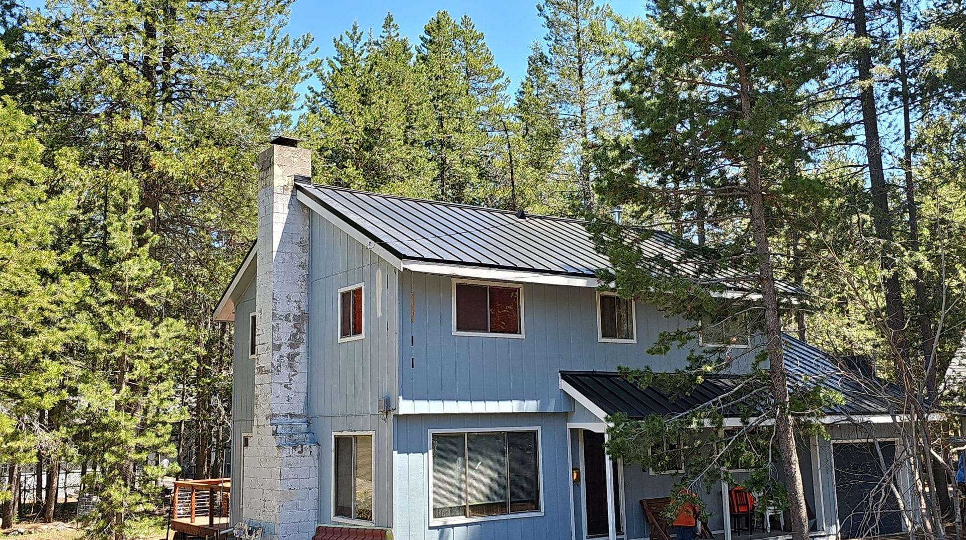 A light blue two-story house with a black metal roof and a stone chimney, surrounded by tall green pine trees on a sunny day.